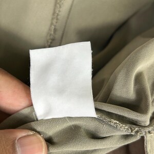 Puede incluir: Primer plano de una etiqueta de ropa blanca en blanco sobre una prenda de color caqui. La etiqueta es rectangular y est&aacute; hecha de una tela blanca lisa. La tela de la prenda es de un color liso y neutro.