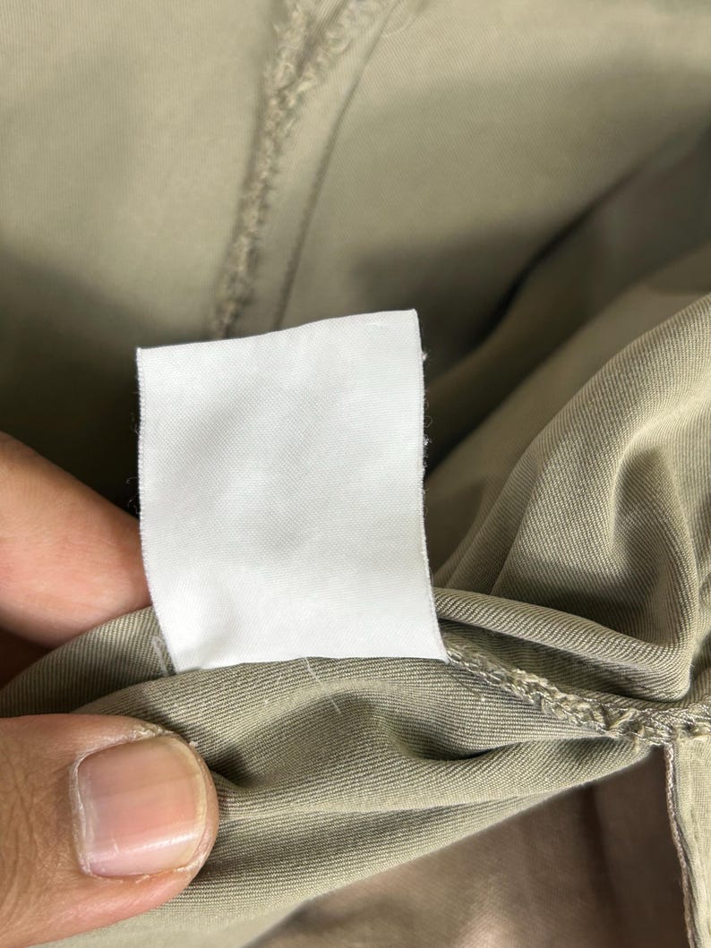 Puede incluir: Primer plano de una etiqueta de ropa blanca en blanco sobre una prenda de color caqui. La etiqueta es rectangular y est&aacute; hecha de una tela blanca lisa. La tela de la prenda es de un color liso y neutro.