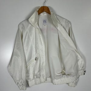 May include: A vintage white jacket with a high collar and a zipper closure. The jacket has a textured, slightly shiny appearance and features a small pocket on the chest. The jacket is hanging on a wooden hanger.