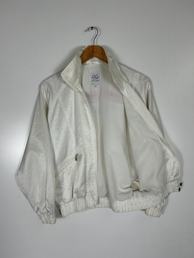 May include: A vintage white jacket with a high collar and a zipper closure. The jacket has a textured, slightly shiny appearance and features a small pocket on the chest. The jacket is hanging on a wooden hanger.