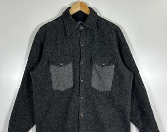 May include: A dark gray wool shirt with two chest pockets and a button-down front. The shirt is hanging on a wooden hanger against a white background. The pockets are a lighter shade of gray.