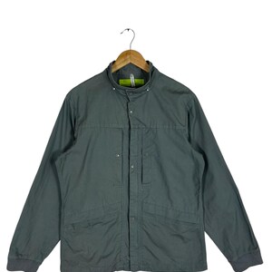 May include: A dark green jacket with a mandarin collar, long sleeves, and a button-up front. The jacket has two front pockets and is hanging on a wooden hanger. The cuffs are a darker shade of green.
