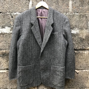 May include: A gray herringbone blazer with two front pockets and a two-button closure. The blazer is hanging on a wooden hanger. The interior lining is a muted purple color. The jacket is made of a textured fabric.