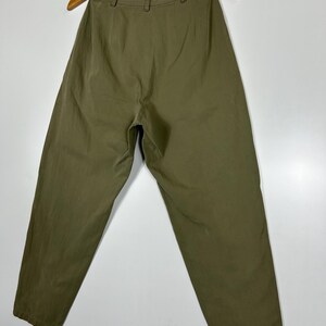 Puede incluir: Pantalones verde oliva colgados de una percha de madera. Los pantalones tienen cintura alta y corte holgado. La tela parece ser un material ligero, adecuado para climas c&aacute;lidos. Los pantalones se muestran sobre un fondo blanco.