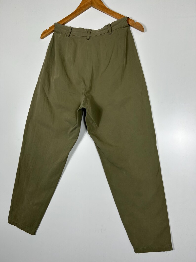 Puede incluir: Pantalones verde oliva colgados de una percha de madera. Los pantalones tienen cintura alta y corte holgado. La tela parece ser un material ligero, adecuado para climas c&aacute;lidos. Los pantalones se muestran sobre un fondo blanco.