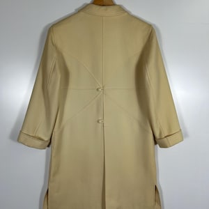 May include: A cream-coloured coat featuring long sleeves with folded cuffs, a collared neckline, and a back panel design with two central buttons. The garment includes side slits at the hem and is displayed on a wooden hanger against a plain background.