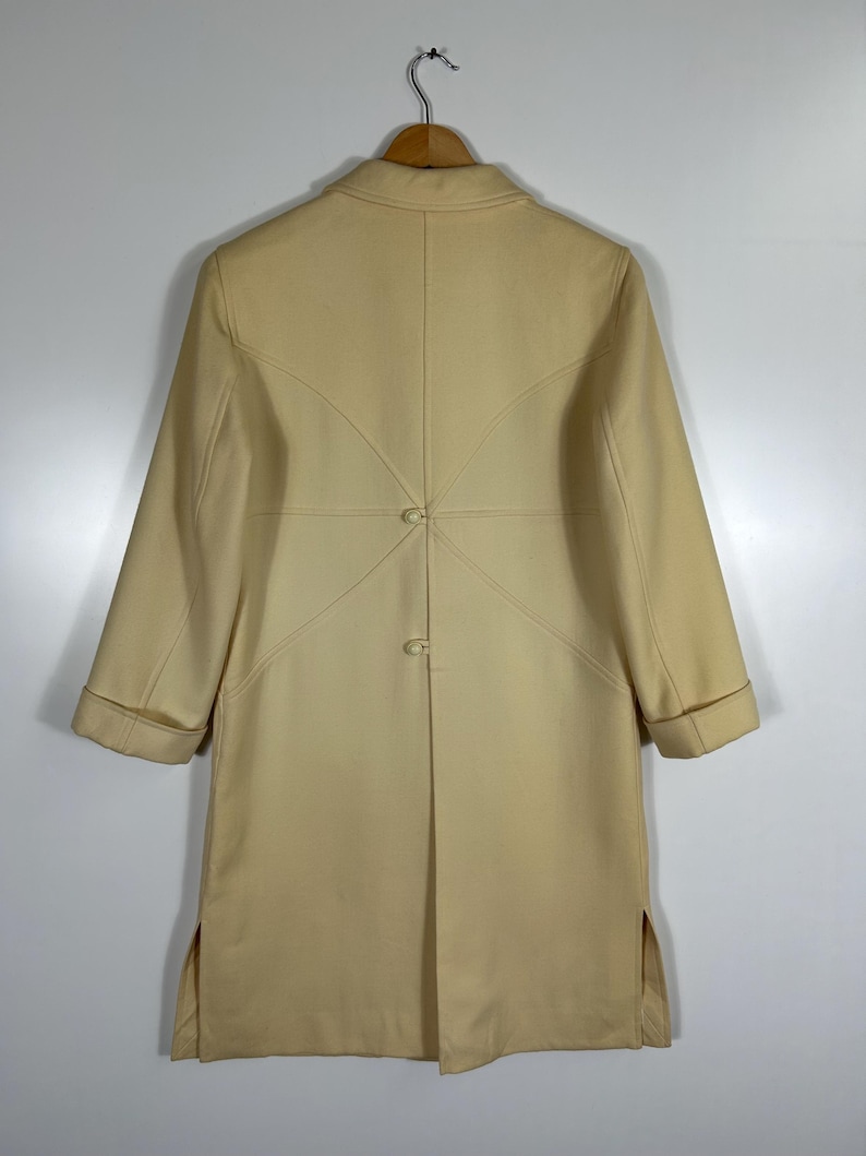 May include: A cream-coloured coat featuring long sleeves with folded cuffs, a collared neckline, and a back panel design with two central buttons. The garment includes side slits at the hem and is displayed on a wooden hanger against a plain background.