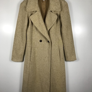 Peut inclure: Un long manteau en tweed beige à double boutonnage. Le manteau présente un col cranté, deux rangées de boutons et des manches longues. Le manteau est suspendu à un cintre en bois.