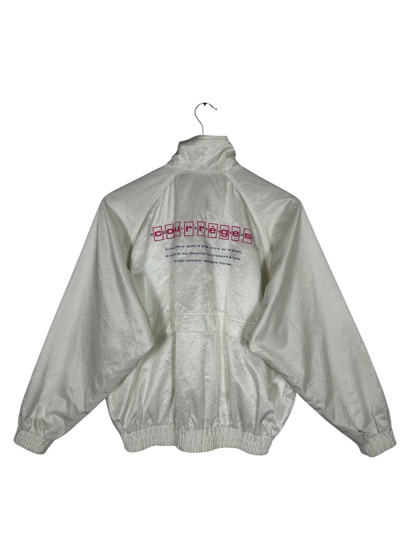 May include: White nylon jacket with a high collar and elastic cuffs. The back of the jacket features the word "courr&egrave;ges" in red block letters above additional text. The jacket has a zipper closure and a gathered waist.