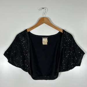 May include: Black sequined bolero jacket with short sleeves, hanging on a wooden hanger. The jacket has a solid black back and a label with text. The bolero is a stylish outerwear piece.