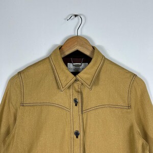 Peut inclure: Une veste jaune moutarde dot&eacute;e d'un col pointu, de manches longues et d'une fermeture boutonn&eacute;e avec des boutons m&eacute;talliques. Le v&ecirc;tement est rehauss&eacute; de surpiq&ucirc;res contrastantes marron fonc&eacute; sur la poitrine et les coutures, avec une &eacute;tiquette Courr&egrave;ges.
