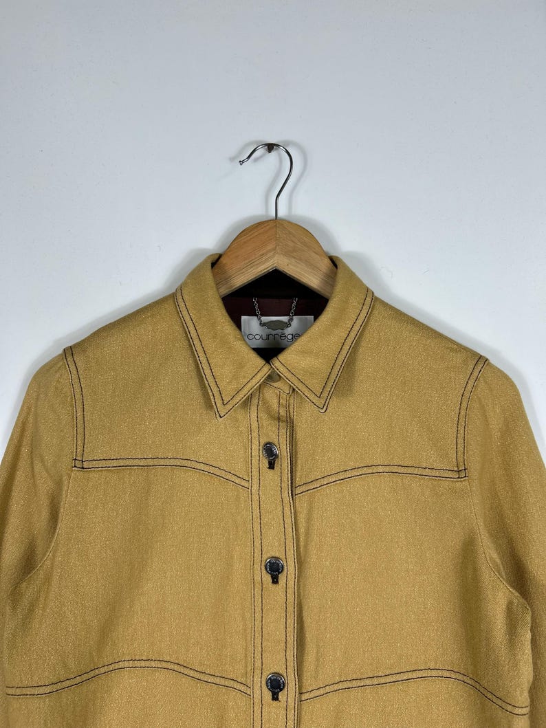 Peut inclure: Une veste jaune moutarde dot&eacute;e d'un col pointu, de manches longues et d'une fermeture boutonn&eacute;e avec des boutons m&eacute;talliques. Le v&ecirc;tement est rehauss&eacute; de surpiq&ucirc;res contrastantes marron fonc&eacute; sur la poitrine et les coutures, avec une &eacute;tiquette Courr&egrave;ges.