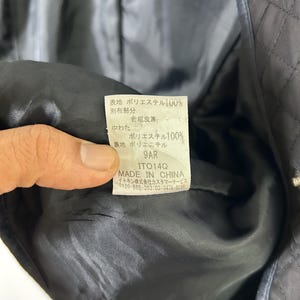 May include: Close-up of a black quilted jacket's interior, revealing a tag with Japanese text and the words "MADE IN CHINA." The tag provides fabric composition details, including 100% polyester for the outer and inner layers. The jacket's lining is black.