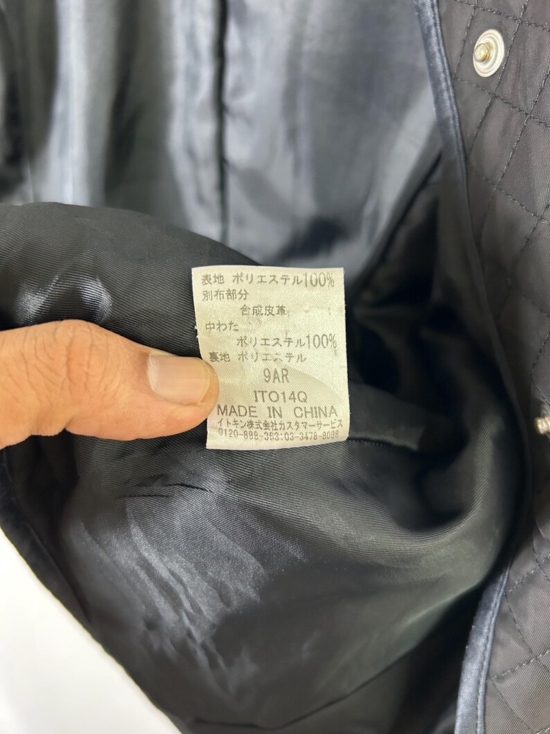 May include: Close-up of a black quilted jacket's interior, revealing a tag with Japanese text and the words "MADE IN CHINA." The tag provides fabric composition details, including 100% polyester for the outer and inner layers. The jacket's lining is black.