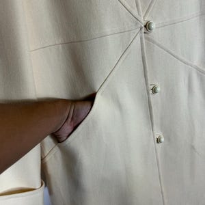 May include: A cream-coloured coat featuring a structured design with diagonal seam detailing radiating from the centre. The garment includes three round, matching cream buttons down the front placket and a deep, functional side pocket.