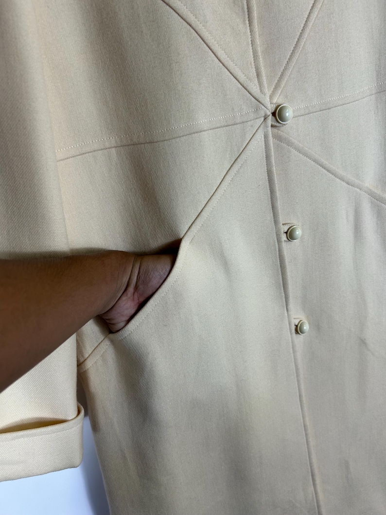 May include: A cream-coloured coat featuring a structured design with diagonal seam detailing radiating from the centre. The garment includes three round, matching cream buttons down the front placket and a deep, functional side pocket.