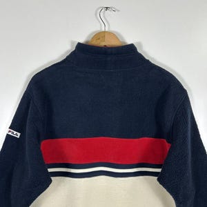 May include: A navy blue, red, and cream-colored fleece pullover with a high collar. The pullover features a red stripe with white accents across the chest and the brand name "FILA" on the sleeve.