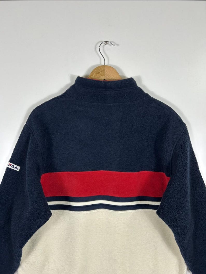 May include: A navy blue, red, and cream-colored fleece pullover with a high collar. The pullover features a red stripe with white accents across the chest and the brand name "FILA" on the sleeve.
