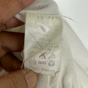 May include: A close-up of a white fabric tag with Japanese text and care symbols. The tag has the text "D-03-46" and "AN 64V". The tag is attached to a white garment.