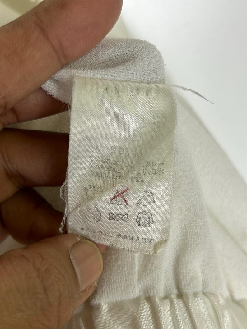 May include: A close-up of a white fabric tag with Japanese text and care symbols. The tag has the text "D-03-46" and "AN 64V". The tag is attached to a white garment.
