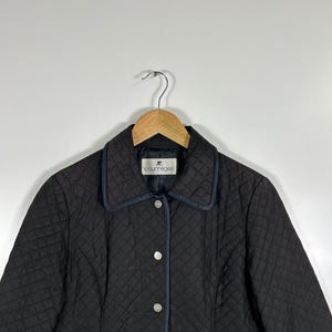 May include: Black quilted jacket with a notched collar and silver-tone buttons. The jacket features a diamond pattern and a small logo on the collar. The jacket is hanging on a wooden hanger.