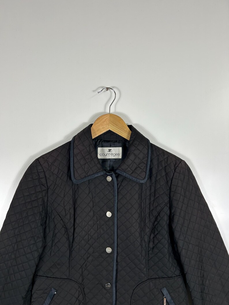 May include: Black quilted jacket with a notched collar and silver-tone buttons. The jacket features a diamond pattern and a small logo on the collar. The jacket is hanging on a wooden hanger.