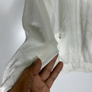 May include: A white, lightweight jacket with a slightly textured surface. The jacket has a simple design with a zipper closure and a gathered hem. The fabric appears to be a thin, possibly synthetic material, suitable for layering.