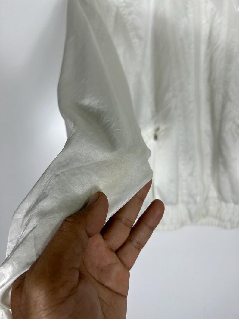 May include: A white, lightweight jacket with a slightly textured surface. The jacket has a simple design with a zipper closure and a gathered hem. The fabric appears to be a thin, possibly synthetic material, suitable for layering.