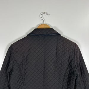 May include: A dark brown quilted jacket with a black leather collar is displayed on a wooden hanger. The jacket has a diamond pattern and a tailored fit. The background is a plain white wall.