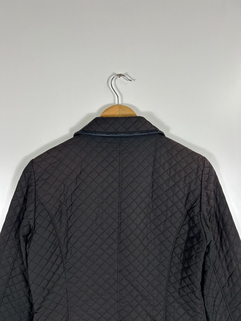 May include: A dark brown quilted jacket with a black leather collar is displayed on a wooden hanger. The jacket has a diamond pattern and a tailored fit. The background is a plain white wall.