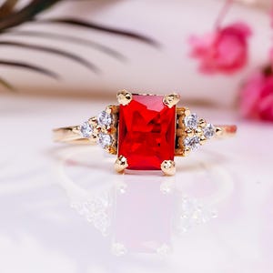 Może przedstawiać: Złoty pierścionek z dużym, prostokątnym, jasno czerwoną kamieniem szlachetnym jako centralnym elementem. Pierścionek jest ozdobiony mniejszymi, przezroczystymi kamieniami szlachetnymi po obu stronach głównego kamienia. Pierścionek jest umieszczony na miękkim, białym tle z różowymi kwiatami.