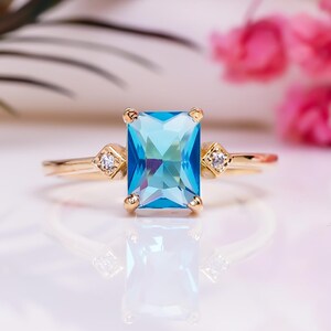 Könnte beinhalten: Ein goldener Ring mit einem rechteckigen, hellblauen Edelstein. Der Ring hat kleine, runde, klare Akzentsteine auf beiden Seiten des Hauptsteins. Der Ring ist vor einem weißen Hintergrund mit rosa Blumen und grünen Blättern platziert.