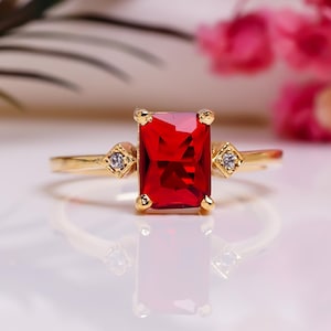Peut inclure: Une bague en or ornée d'une grande pierre rouge rectangulaire au centre. La bague est ornée de petites pierres d'accent claires de chaque côté de la pierre principale. Le fond est flou, avec des fleurs roses.