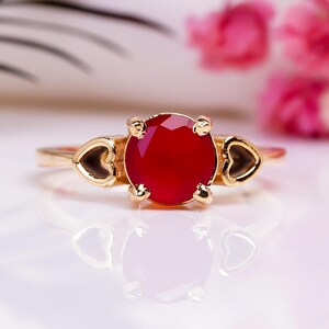 Puede incluir: Un anillo de oro con una gran gema roja redonda. El anillo presenta detalles en forma de corazón a los lados de la gema. El anillo está sobre un fondo blanco suave con flores rosas y hojas verdes.