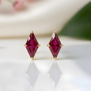 Könnte beinhalten: Ein Paar goldene Ohrstecker mit rautenförmigen, tiefroten Edelsteinen. Die Ohrringe sind vor einem weißen Hintergrund mit zarten rosa Blüten und einem grünen Blatt im Hintergrund platziert. Die Ohrringe spiegeln sich auf der weißen Oberfläche.