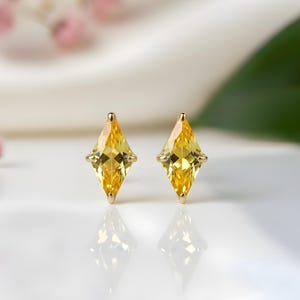 Puede incluir: Un par de pendientes de oro con gemas amarillas en forma de diamante. Los pendientes están engastados en oro. El fondo está borroso, con toques de flores rosas y una superficie blanca. Reflejan la luz.