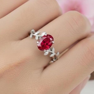 Può includere: Anello d'argento con una gemma rosso scuro ovale. La fascia dell'anello presenta un delicato design a foglie su entrambi i lati della gemma, creando un'estetica elegante e ispirata alla natura. Un gioiello.