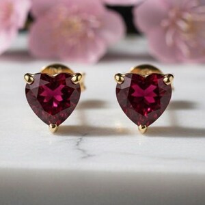 Pode incluir: Um par de brincos de ouro com pedras preciosas vermelhas escuras facetadas em forma de coração. Os brincos estão colocados sobre uma superfície branca, com flores rosa desfocadas no fundo. Um item de joalheria clássico.