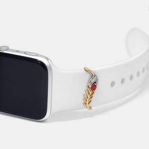 Könnte beinhalten: Ein weißes Apple Watch Armband mit einem goldenen Feder-Anhänger. Der Feder-Anhänger hat einen roten Edelstein und klare Strasssteine.