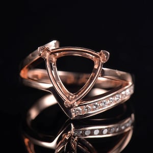 May include: Rose gold ring with a triangular setting and a band adorned with small, clear gemstones. The ring's design features a split shank and a polished finish, reflecting light against a dark background.