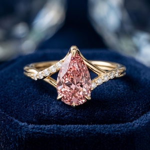 Può includere: Un anello d'oro con una gemma rosa a forma di pera. La fascia presenta un design delicato con piccole pietre scintillanti. L'anello è appoggiato su una superficie di velluto blu scuro.