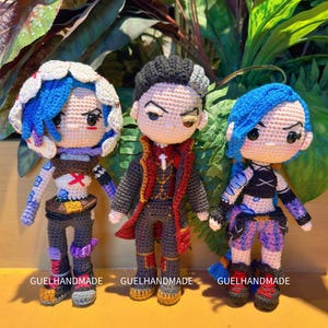 May include: Three handmade crochet dolls with detailed outfits and hairstyles. The dolls feature blue, black, and brown hair, and are dressed in various outfits. The dolls are standing in front of a backdrop of green foliage. The text "GUELHANDMADE" is visible on the base of each doll.