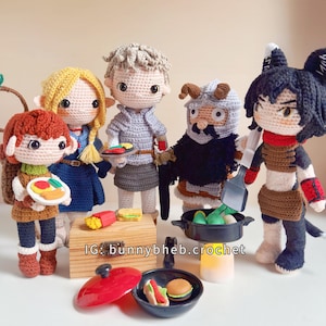 May include: Five crocheted dolls with yarn hair and detailed outfits stand around a wooden box and miniature cooking accessories. The dolls hold small food items. A black frying pan contains a toy burger and hot dog. Text reads IG: bunnybheb.crochet.