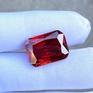 May include: A faceted, rectangular-cut, deep red gemstone. The gem has multiple facets that reflect light, creating a brilliant sparkle. The gemstone is displayed on a white surface.