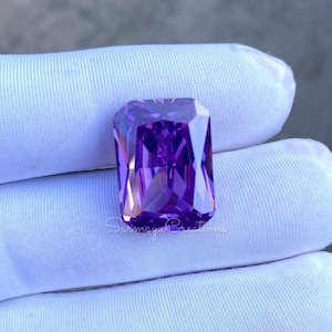 May include: A large, rectangular-cut, faceted purple gemstone. The gem is held on a white fabric surface. The gem has a brilliant, reflective surface, showcasing its internal structure. The image is well-lit, highlighting the gem's color and clarity.