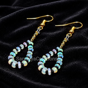 May include: A pair of drop earrings featuring a teardrop-shaped loop of alternating iridescent white opal-like beads and small black beads. The earrings are suspended from gold-toned metal hooks by a short chain of gold-toned metal beads.