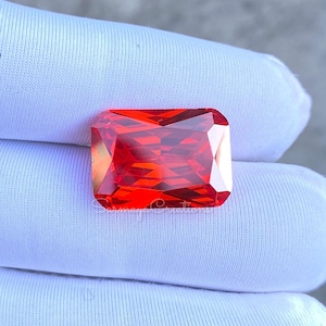 May include: A vibrant, rectangular-cut gemstone with a deep, saturated orange-red hue and multifaceted surface that reflects light. The stone is positioned against a white fabric background, with the text "Sohmaya Creations" visible below it.