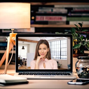 Könnte beinhalten: Eine Frau in einem Videogespräch auf einem Laptop in einem modernen Büro. Sie trägt eine rosa Bluse und hat die Hand am Kinn. Der Laptop steht auf einem Schreibtisch mit einer Lampe, einem Notizbuch und einer Pflanze.