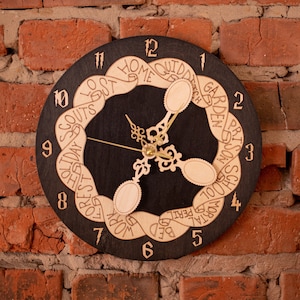Puede incluir: Un reloj de madera negro con un diseño único que presenta una cinta con palabras como "Home", "Work", "Bed" y "School" alrededor de la esfera del reloj. Las manecillas del reloj son doradas y los números son blancos.