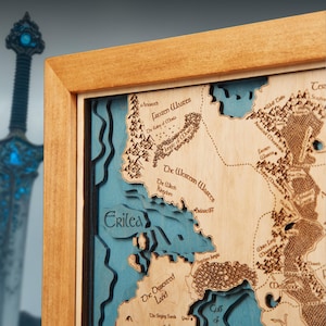 May include: A wooden map with a detailed laser-cut design, featuring a fantasy world with geographical features and text labels. The map is framed in light brown wood, with blue layers. A decorative sword is visible in the background.